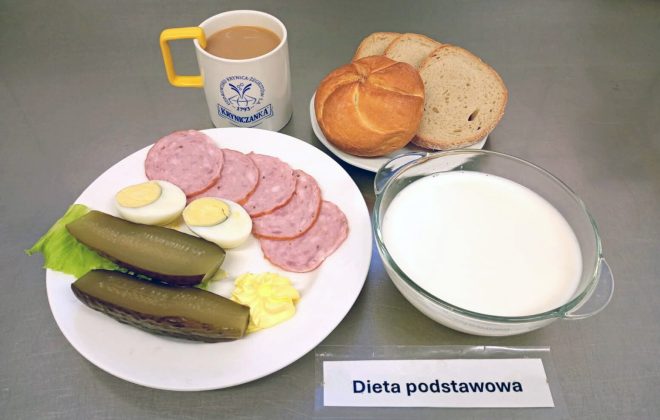 Śniadanie w diecie podstawowej: talerz z plasterkami kiełbasy, jajkiem na twardo, ogórkiem kiszonym, sałatą i porcją masła. Obok talerz z trzema kromkami chleba i bułką. Do posiłku podano kubek z kawą z mlekiem oraz miseczkę z mlekiem lub jogurtem naturalnym