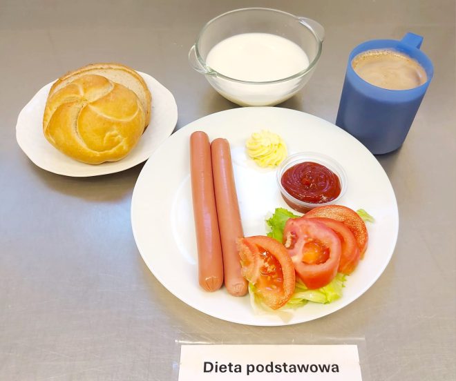 Na talerzu ułożono dwa parówki, kilka plastrów pomidora z sałatą, porcję masła oraz ketchup w małym pojemniczku. Obok znajduje się bułka kajzerka z kromką chleba pszennego, miseczka z mlekiem i kubek z kawą z mlekiem w niebieskim kubku. Przed zestawem leży karteczka z napisem „Dieta podstawowa”