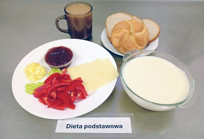 Na zdjęciu widoczny jest talerz z plasterkami żółtego sera, kawałkami czerwonej papryki, sałatą, porcją masła i dżemem w małym pojemniczku. Obok ustawiono talerz z pieczywem (kajzerka i kromki chleba), miseczkę z kaszą manną na mleku oraz kubek kawy z mlekiem. Przed zestawem leży karteczka z napisem „Dieta podstawowa”