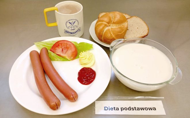 Na talerzu dwa parówki z dodatkiem liścia sałaty i plastra pomidora, obok porcja musztardy i ketchupu. W tle kajzerka z kromkami chleba, miseczka z mlekiem oraz kubek kawy z mlekiem. Na stoliku przed talerzem leży kartka z napisem „Dieta podstawowa”