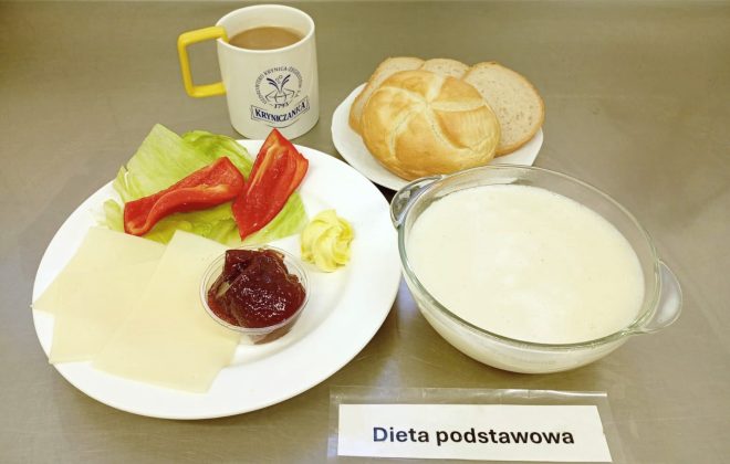 Na talerzu dwa plastry sera żółtego, liść sałaty, kawałek papryki czerwonej oraz porcja dżemu w małym pojemniczku. Obok pieczywo: kajzerka i kromki chleba, miseczka z mlekiem oraz kubek kawy z mlekiem. Na pierwszym planie umieszczona jest kartka z napisem „Dieta podstawowa”