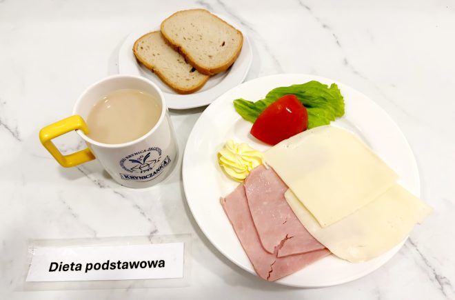 Na białym talerzu znajdują się dwa plasterki szynki, dwa plastry sera żółtego, kawałek pomidora, liść sałaty i porcja masła. Obok talerz z kromkami chleba oraz kubek kawy z mlekiem. Na stoliku kartka z napisem „Dieta podstawowa”