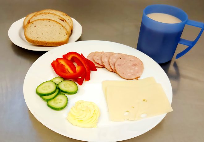 Śniadanie składające się z talerza z plasterkami wędliny, sera żółtego, masła, ogórka i papryki. Obok leży talerzyk z kromkami chleba oraz kubek z kawą z mlekiem lub herbatą z mlekiem