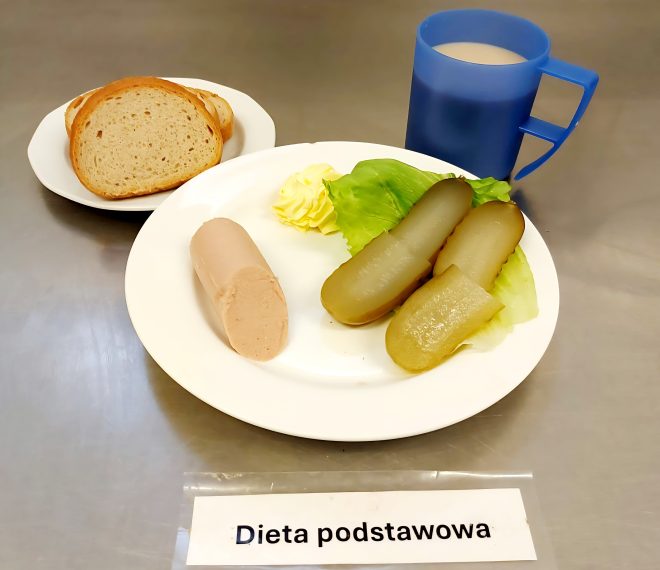 Talerz z plasterkiem pasztetowej, dwoma kawałkami ogórka kiszonego, liściem sałaty i porcją masła. Obok kromki chleba na talerzyku oraz kubek z kawą z mlekiem. Na stole znajduje się karteczka „Dieta podstawowa”