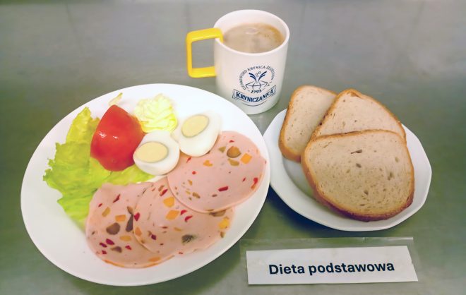 Posiłek w diecie podstawowej: talerz z plasterkami wędliny drobiowej z warzywami, połową pomidora, listkiem sałaty, dwoma jajkami na twardo i porcją masła. Obok znajduje się talerzyk z trzema kromkami pieczywa pszennego oraz kubek z kawą zbożową lub herbatą