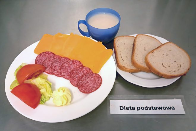 Zestaw śniadaniowy w diecie podstawowej: trzy kromki chleba, kilka plastrów salami, plastry sera żółtego, sałatka z pomidora i sałaty, porcja masła oraz kawa z mlekiem w niebieskim kubku