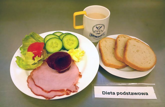 Zestaw śniadaniowy w diecie podstawowej: trzy kromki chleba pszennego, plasterki szynki, sałatka z pomidora, ogórka i sałaty, porcja masła, kawa z mlekiem podana w kubku „Kryniczanka”