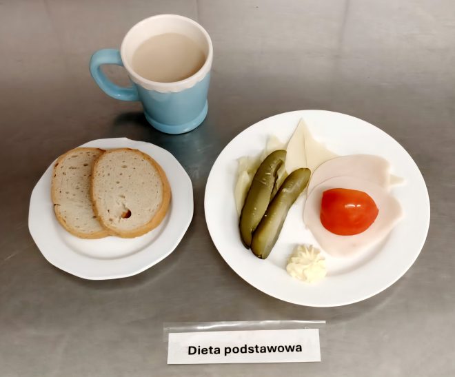 Śniadanie w ramach diety podstawowej. Na talerzu ułożono plaster sera żółtego, dwa plastry chudej wędliny drobiowej, kawałek pomidora, ogórki kiszone oraz porcję masła. Obok znajduje się talerzyk z dwiema kromkami chleba. Do posiłku dołączono kubek kawy z mlekiem