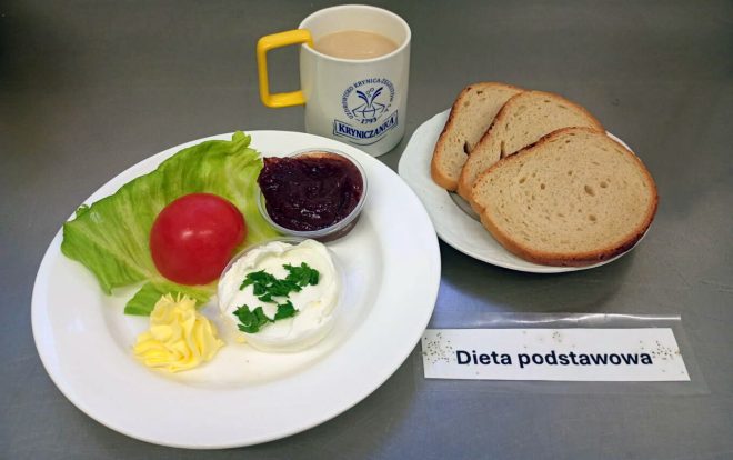 Na stole znajduje się zestaw śniadaniowy opisany jako „Dieta podstawowa”. Na białym talerzu ułożono liść sałaty z całym pomidorem, porcję twarożku posypanego natką pietruszki, porcję masła oraz mały kubeczek z dżemem. Obok podano trzy kromki chleba pszennego oraz kubek kawy z mlekiem w filiżance z żółtym uchem i logo „Kryniczanka”