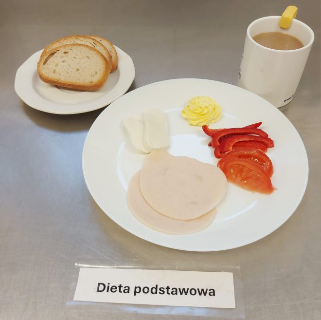 Talerz z dwoma plasterkami wędliny drobiowej, plastrem pomidora, kilkoma paskami papryki czerwonej, kawałkiem sera białego i porcją masła. Obok kromki chleba pszennego na osobnym talerzyku oraz kubek z kawą z mlekiem. Pod talerzem widoczny napis „Dieta podstawowa”