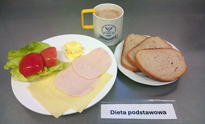 Śniadanie w diecie podstawowej: kromki chleba pszennego, plasterki sera żółtego i wędliny drobiowej, liść sałaty z kawałkami pomidora, porcja masła oraz kubek kawy z mlekiem
