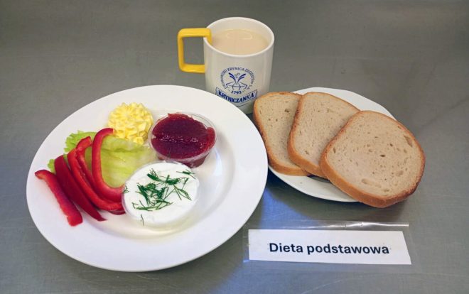 Na tacy podano posiłek oznaczony jako „Dieta podstawowa”. Na talerzu znajdują się kromki chleba pszennego i bułka, a obok talerza miska z jasną zupą mleczną. Na drugim talerzyku leżą plasterki papryki, liść sałaty, porcje twarożku ze szczypiorkiem, masła i dżemu. Do posiłku podano kubek z napojem