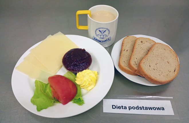 Na zdjęciu śniadanie w diecie podstawowej. Na białym talerzu ułożono kilka plastrów sera żółtego, porcję sałaty, kawałek pomidora, porcję masła oraz małą miseczkę z konfiturą. Obok znajduje się talerzyk z trzema kromkami chleba pszennego. Do zestawu podano kubek z kawą z mlekiem