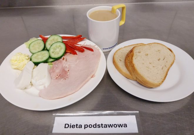 Śniadanie w diecie podstawowej: talerz z wędliną drobiową, plastrami ogórka i czerwonej papryki, porcją twarogu i masła; obok drugi talerz z kromkami jasnego pieczywa. Do posiłku podany biały kubek z kawą z mlekiem