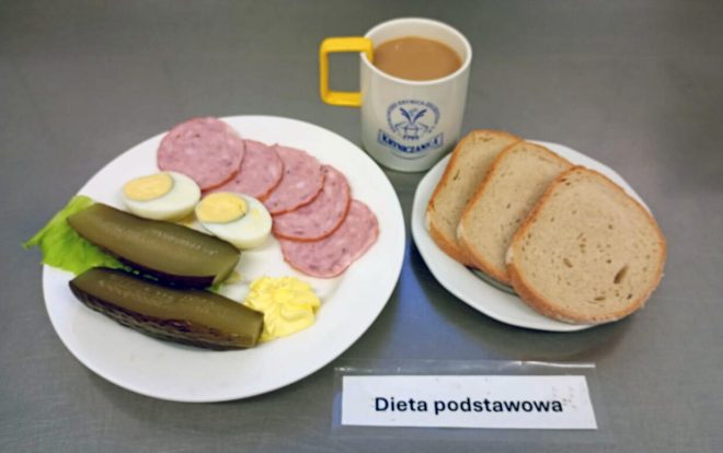 Śniadanie w diecie podstawowej: talerz z plasterkami wędliny, jajkiem na twardo, ogórkiem kiszonym i porcją masła. Obok drugi talerz z trzema kromkami chleba pszenno-żytniego. Do posiłku podano kubek z kawą z mlekiem