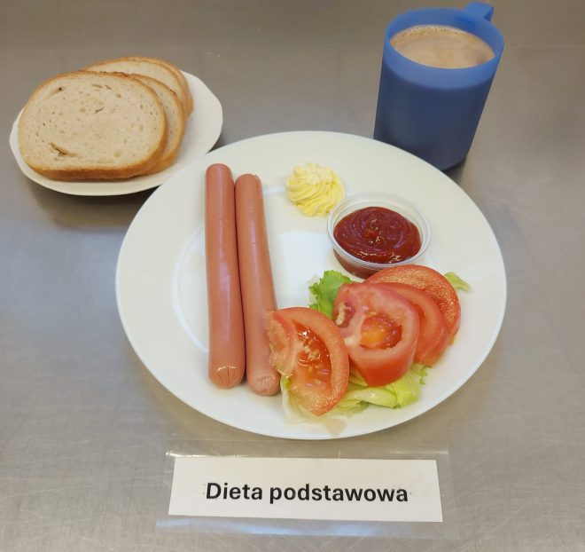 Na zdjęciu przedstawiony jest zestaw śniadaniowy oznaczony etykietą „Dieta podstawowa”. Na białym talerzu znajdują się dwie parówki podane z plasterkami świeżego pomidora ułożonymi na liściach sałaty, porcją masła w rozetce oraz sosem pomidorowym w małym pojemniczku. Obok na osobnym talerzyku ułożono kilka kromek pieczywa pszennego. Do posiłku podano także kubek z gorącym napojem, prawdopodobnie kawą z mlekiem lub kakao, w niebieskim naczyniu