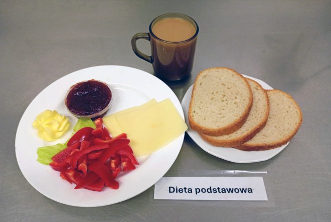 Na zdjęciu widoczny jest zestaw składający się z trzech kromek chleba pszennego, plastrów żółtego sera, masła, sałaty i kawałków czerwonej papryki oraz porcji dżemu. Obok stoi szklanka kawy z mlekiem. Na stoliku położono kartkę z napisem „Dieta podstawowa”