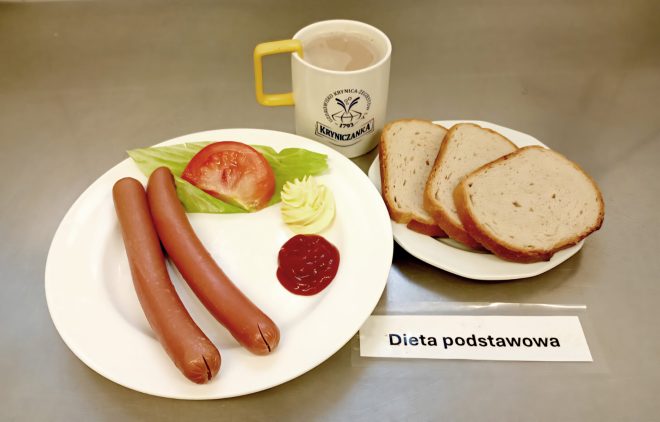 Talerz z dwoma parówkami, kawałkiem pomidora, liściem sałaty, porcją masła i ketchupem. Obok talerzyk z trzema kromkami chleba oraz kubek z kawą z mlekiem. Na stole karteczka z napisem „Dieta podstawowa”