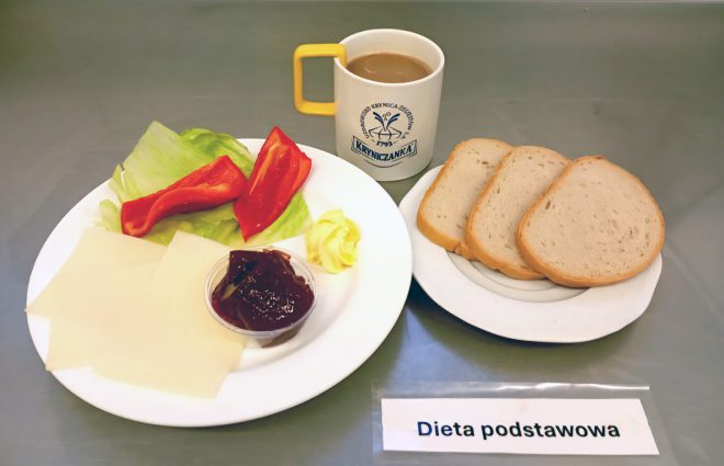 Na tacy ustawiono zestaw śniadaniowy. Na talerzu znajdują się trzy kromki chleba pszennego, a obok plasterki sera żółtego, kawałek sałaty, paski czerwonej papryki, mała porcja dżemu oraz kostka masła. Do posiłku podano kubek kawy z mlekiem. Na tacy umieszczono etykietę „Dieta podstawowa”
