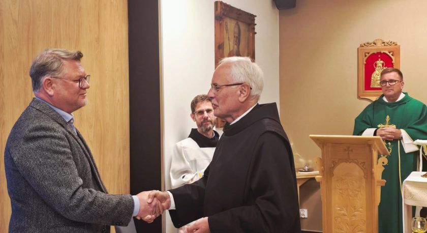 W kaplicy zakonnik w habicie ściska dłoń mężczyzny w marynarce. W tle dwóch duchownych w zielonych szatach liturgicznych, obok ołtarz i obraz Matki Bożej