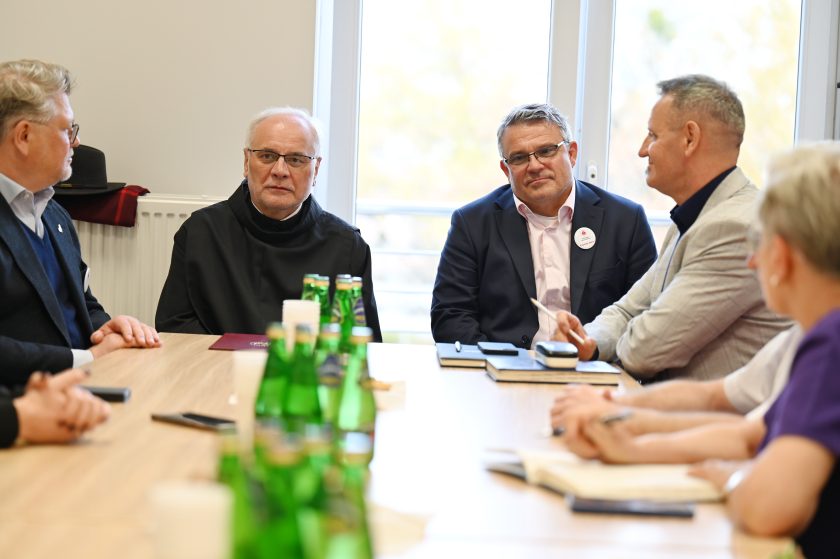 Przy stole w sali konferencyjnej siedzą uczestnicy spotkania – zakonnik w czarnym habicie oraz kilku mężczyzn w garniturach. Na stole woda mineralna, notatniki i dokumenty. Rozmowa przebiega w skupieniu