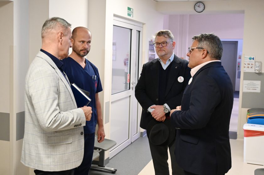 Czterech mężczyzn rozmawia w szpitalnym korytarzu. Jeden z nich ubrany jest w granatowy strój medyczny, pozostali w garniturach i marynarkach. Spotkanie odbywa się w spokojnej atmosferze, przy drzwiach prowadzących do części klinicznej