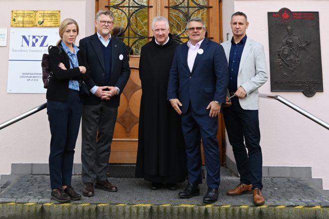 Grupa pięciu osób – kobieta, trzej mężczyźni w garniturach i zakonnik w habicie – stoi przed wejściem do budynku Bonifraterskiego Centrum Medycznego. Obok widoczne tablice informacyjne z logo NFZ i pamiątkowa płaskorzeźba