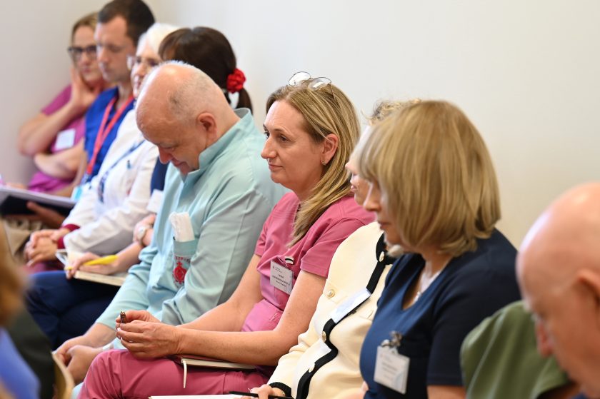 Grupa uczestników spotkania siedzi w jednym rzędzie w sali konferencyjnej. Widać personel medyczny w kolorowych uniformach oraz osoby w eleganckich strojach. Wszyscy skupieni na przebiegu spotkania