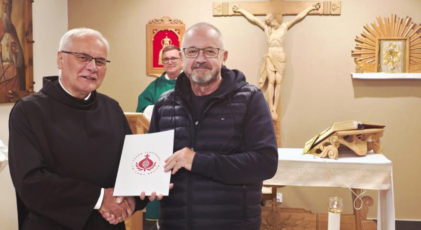 W kaplicy zakonnik w habicie wręcza dokument z logo Zakonu Bonifratrów mężczyźnie w czarnej kurtce. Obaj stoją przed ołtarzem z krzyżem i otwartym Pismem Świętym
