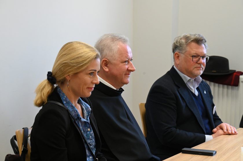 Kobieta, zakonnik i mężczyzna w garniturze siedzą obok siebie przy stole w sali spotkań. Wszyscy uśmiechnięci, w tle widać białą ścianę i okno z wpadającym światłem dziennym