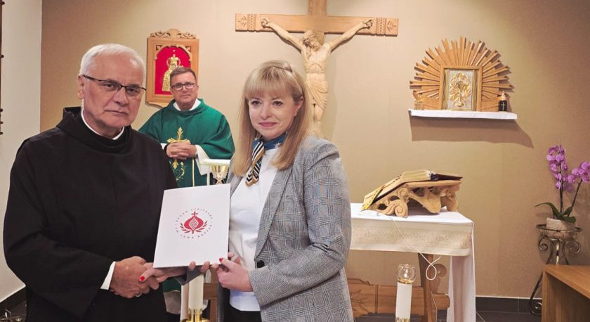 Zakonnik w habicie i kobieta w szarej marynarce stoją przed ołtarzem. Kobieta trzyma teczkę z logo Zakonu Bonifratrów. W tle kapłan w zielonej albie i drewniany krucyfiks