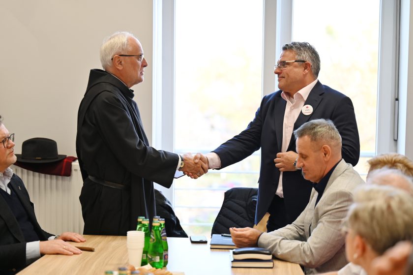 Zakonnik w habicie i mężczyzna w garniturze podają sobie dłonie na zakończenie spotkania przy stole. Pozostali uczestnicy siedzą obok, a na stole stoją butelki z wodą i notatniki