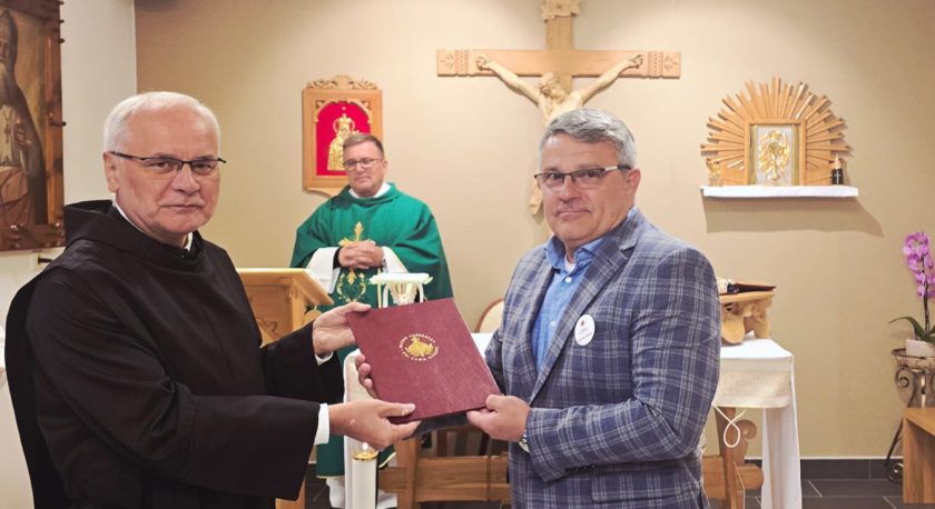 Zakonnik w czarnym habicie wręcza bordową teczkę z godłem Zakonu Bonifratrów mężczyźnie w niebieskiej koszuli i kratkowanej marynarce. Za nimi ołtarz z krucyfiksem