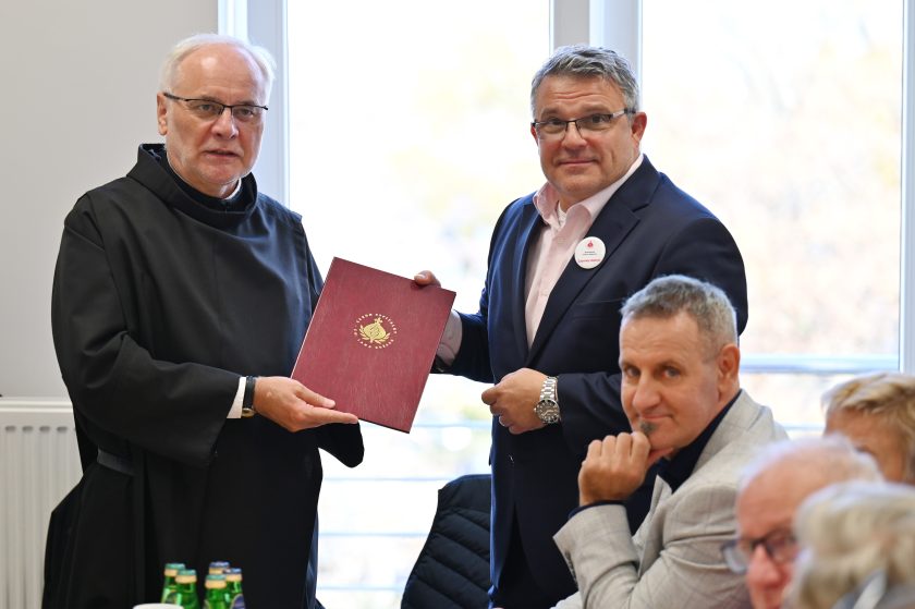 Zakonnik w czarnym habicie i mężczyzna w garniturze stoją obok siebie, trzymając wspólnie bordową teczkę z godłem Zakonu Bonifratrów. Wokół stołu pozostali uczestnicy spotkania w rozmowie