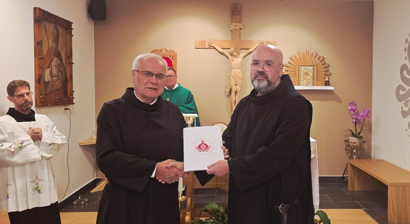 Dwóch zakonników w habitach stoi przed ołtarzem. Jeden z nich wręcza drugiemu dokument z logo Zakonu Bonifratrów. W tle obraz świętego i kapłan w zielonej albie przy ołtarzu