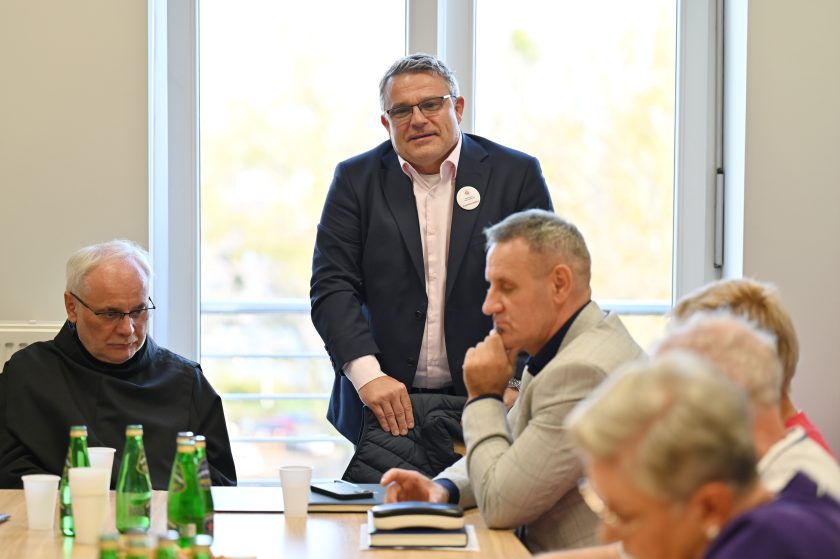 Mężczyzna w garniturze z przypiętą plakietką Bonifraterskiego Centrum Medycznego stoi przy stole, zwracając się do uczestników spotkania. Obok zakonnik i inni słuchacze w skupieniu notują