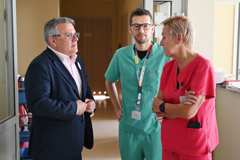 W szpitalnym korytarzu mężczyzna w garniturze rozmawia z dwojgiem pracowników medycznych – kobietą w czerwonym i mężczyzną w zielonym uniformie. Dyskusja dotyczy prawdopodobnie kwestii organizacyjnych