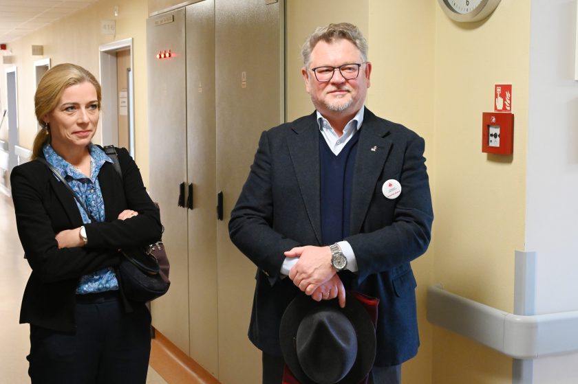 Kobieta w czarnej marynarce i mężczyzna w garniturze stoją w korytarzu szpitalnym. Oboje z przypiętymi plakietkami Bonifraterskiego Centrum Medycznego, w tle drzwi do sal chorych
