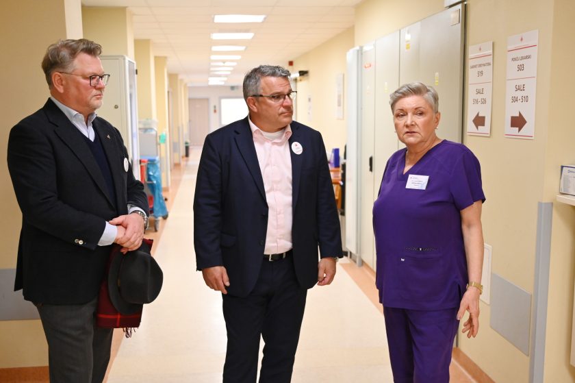 Dwoje przedstawicieli Bonifraterskiego Centrum Medycznego rozmawia z kobietą w fioletowym stroju medycznym w szpitalnym korytarzu. Na ścianie widoczne są tabliczki z oznaczeniami gabinetów i sal