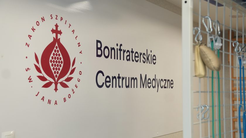 Logo Zakonu Szpitalnego św. Jana Bożego i napis „Bonifraterskie Centrum Medyczne” na ścianie w sali rehabilitacyjnej