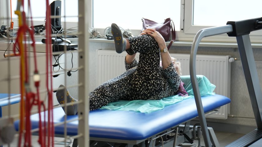 Pacjentka wykonuje ćwiczenia rozciągające na leżance rehabilitacyjnej. Leży na plecach i unosi zgiętą nogę w kierunku klatki piersiowej