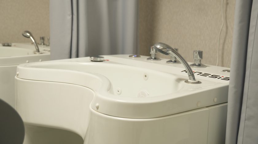 Zbliżenie na biały sprzęt do hydroterapii z kranami i pokrętłami. Na urządzeniu widoczny napis „Kinesis”