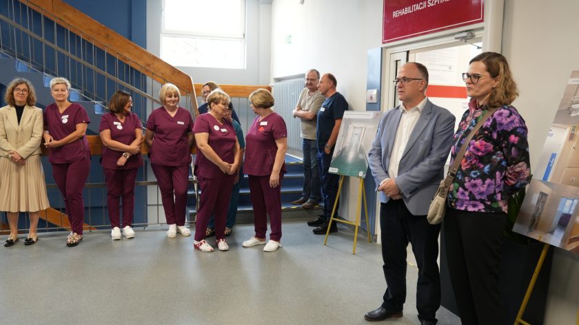 Grupa pracowników Bonifraterskiego Centrum Medycznego oraz gości stoi w holu przy wejściu do działu rehabilitacji. Kobiety w bordowych uniformach rozmawiają, obok nich stoją osoby w eleganckich strojach i kilku mężczyzn pod ścianą