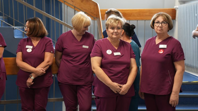 Grupa pielęgniarek w bordowych uniformach stoi na tle niebieskiej klatki schodowej, rozmawiając i uśmiechając się podczas spotkania w Bonifraterskim Centrum Medyczny
