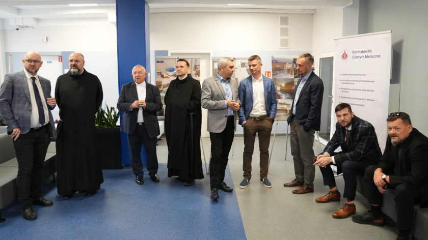 Grupa mężczyzn, wśród nich zakonnicy i przedstawiciele władz Bonifraterskiego Centrum Medycznego, stoi w sali z banerami przedstawiającymi modernizację szpitala