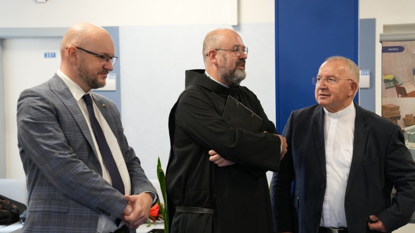 Trzech uczestników spotkania — zakonnik w czarnej habicie i dwóch mężczyzn w garniturach — prowadzi rozmowę w korytarzu Bonifraterskiego Centrum Medycznego