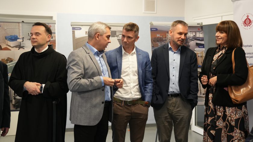 Uczestnicy spotkania rozmawiają w holu szpitala. Wśród nich mężczyźni w garniturach, zakonnik oraz kobieta w ciemnej sukience w wzory. W tle plansze ze zdjęciami z remontu placówki