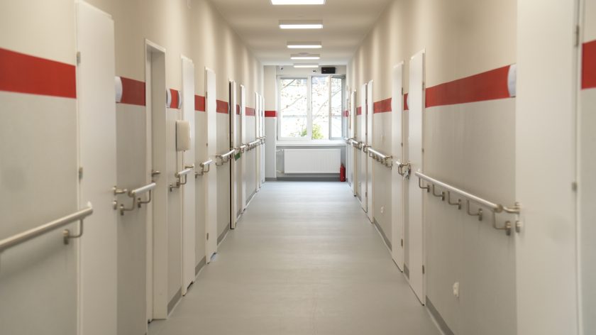 Nowo wyremontowany korytarz Bonifraterskiego Centrum Medycznego. Jasne ściany z czerwonym pasem, poręcze wzdłuż ścian i drzwi do sal pacjentów prowadzące do okna na końcu korytarza