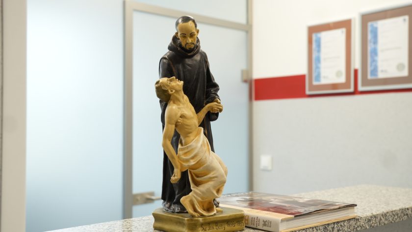 Figura św. Jana Bożego trzymającego chorego, ustawiona na blacie recepcji w Bonifraterskim Centrum Medycznym. W tle widać fragment ściany z certyfikatami i drzwi do gabinetu