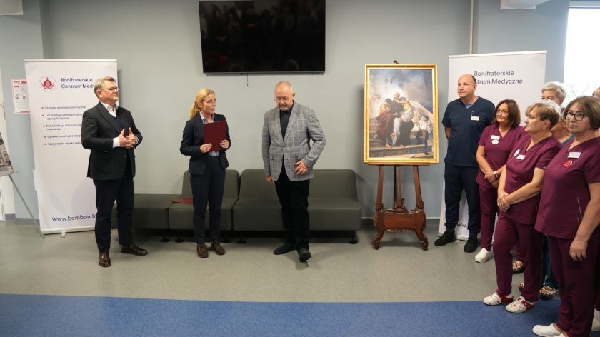 Trzy osoby — kobieta z teczką, mężczyzna w marynarce w kratę i kolejny w garniturze — stoją przy obrazie św. Jana Bożego podczas uroczystości w Bonifraterskim Centrum Medycznym. Obok nich personel medyczny w bordowych strojach